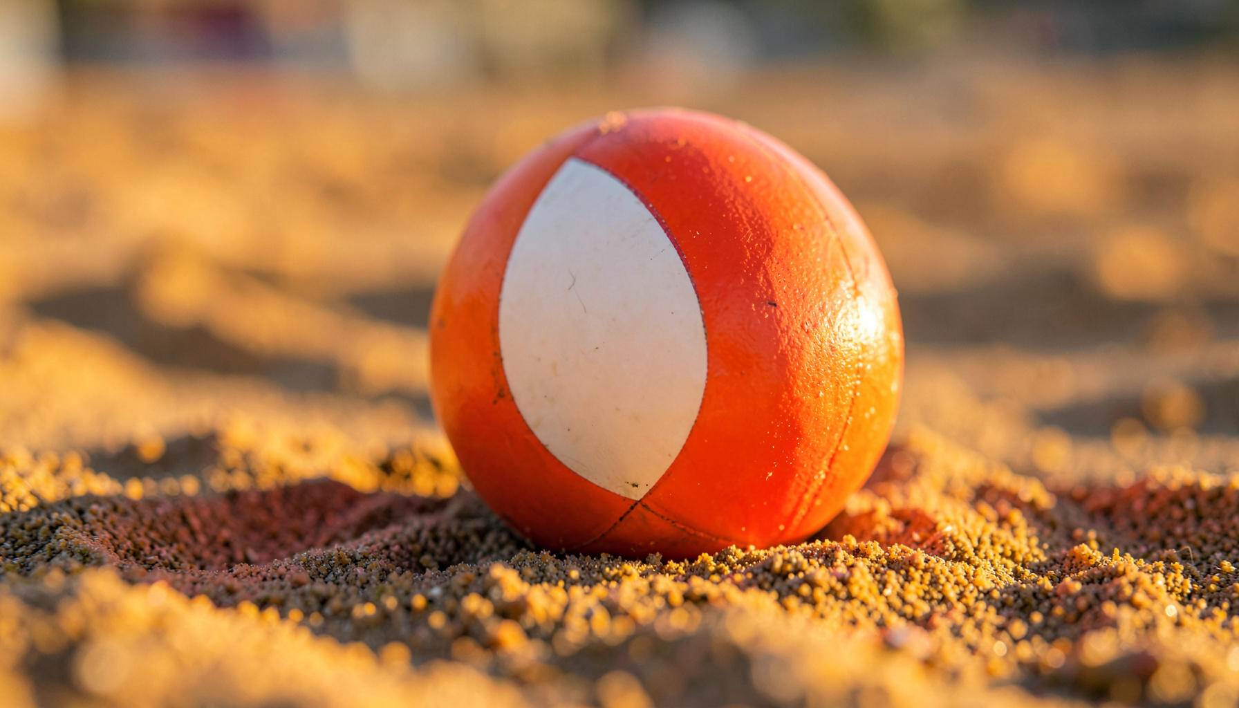 Plaj voleybolu filesinin hemen önünde, kumda duran turuncu-beyaz bir voleybol topunun yakın çekimi; yumuşak akşam ışığı, sıcak kum tonları